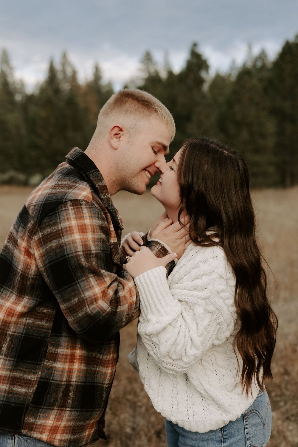 A romantic couple in autumn attire sharing tender kisses in a field with trees in the background.