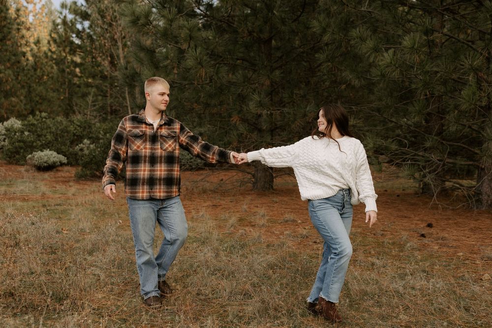 A couple in casual fall attire dances together in a wooded outdoor setting during golden hour.