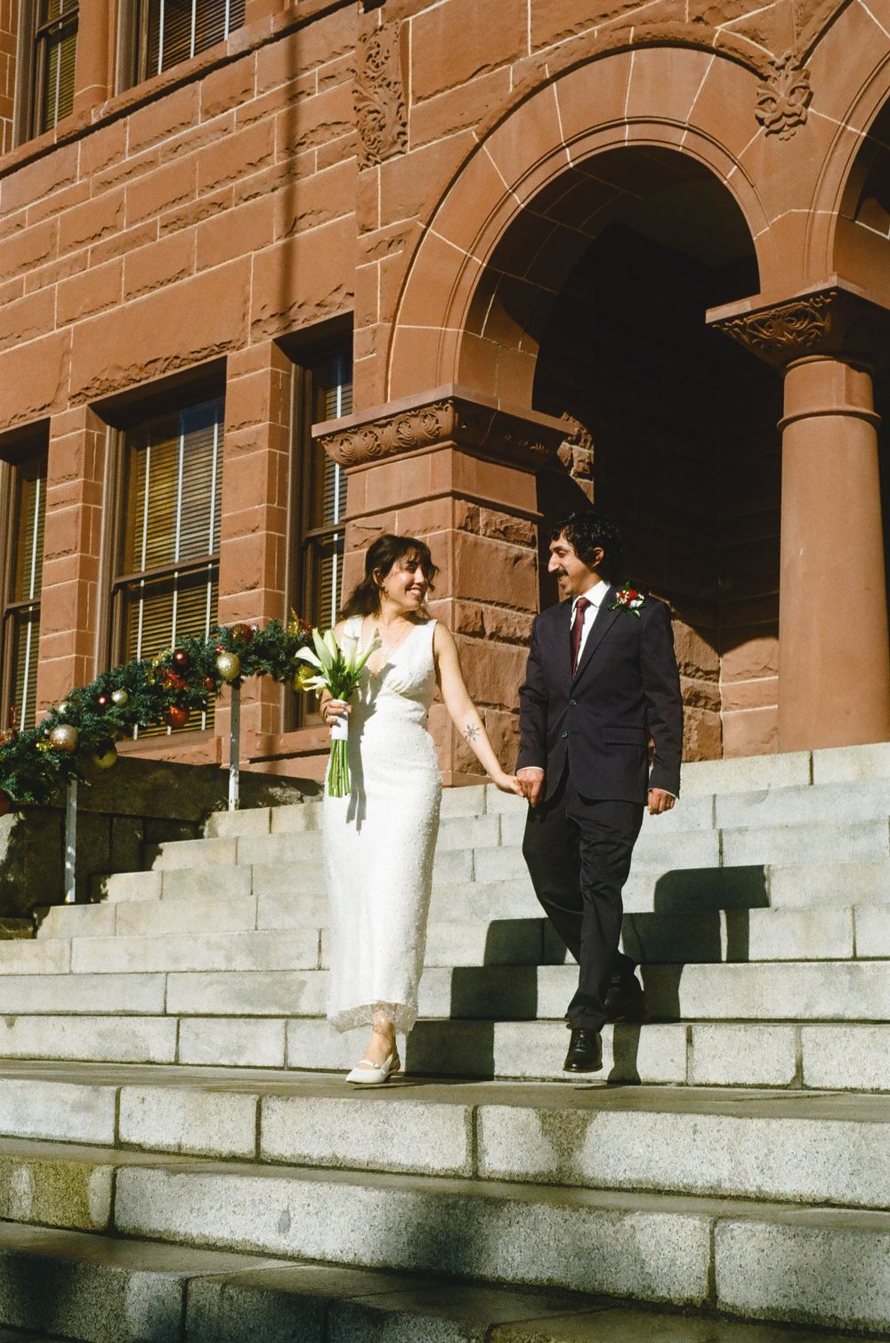 Newlyweds descending steps of a historic red sandstone building, bride holding white bouquet.