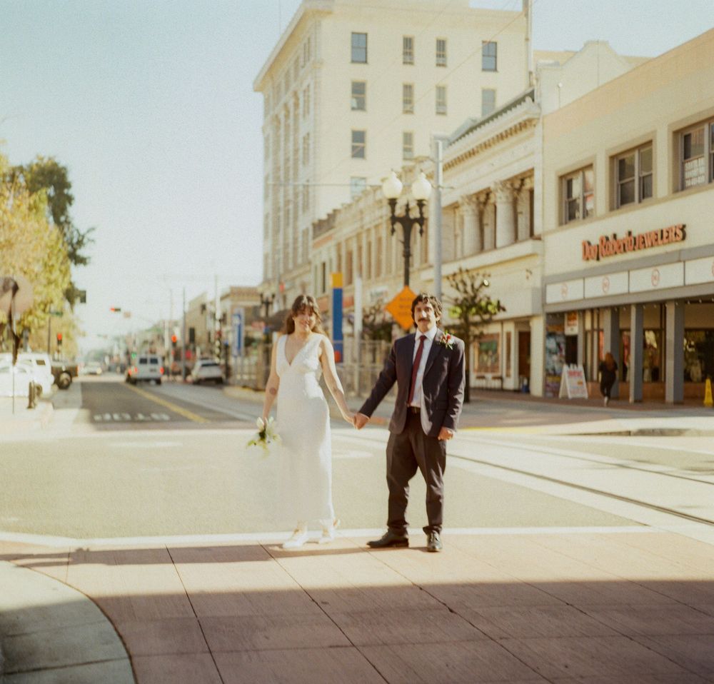 Bride and groom holding hands while crossing a sunny downtown street lined with shops.