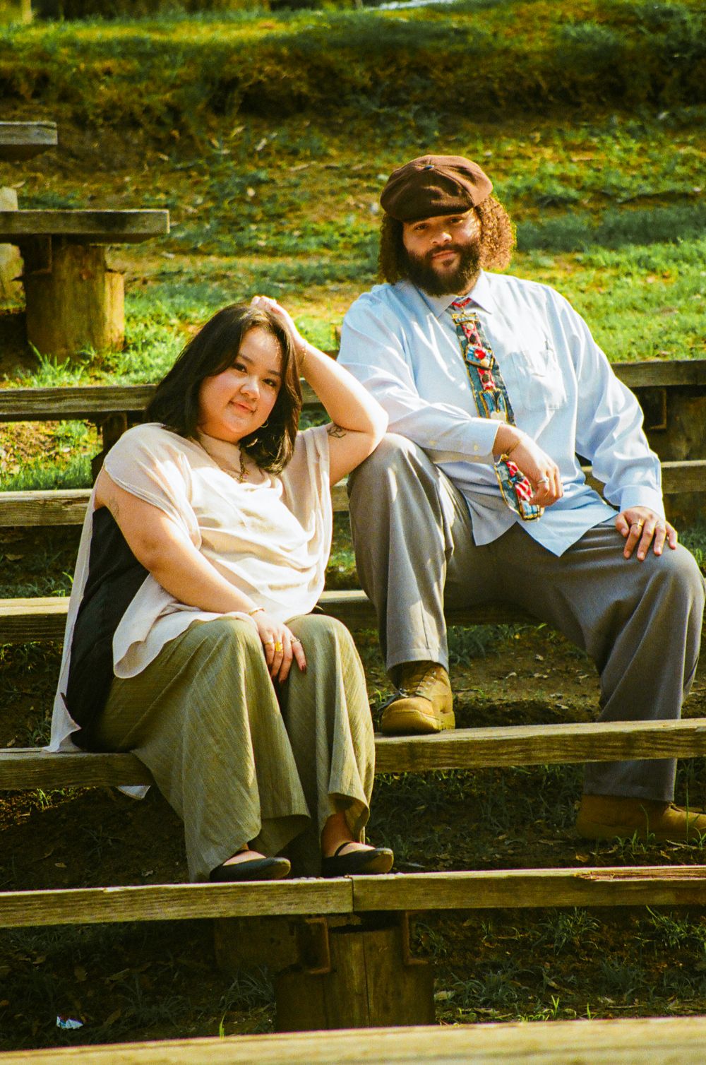 Couple sitting on wooden bleachers outdoors in golden sunlight surrounded by green hills.