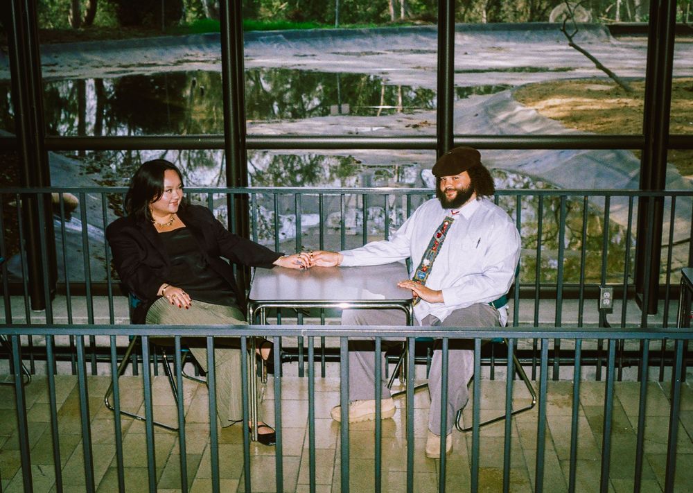 Couple holding hands across a table by large windows overlooking a lush green landscape.