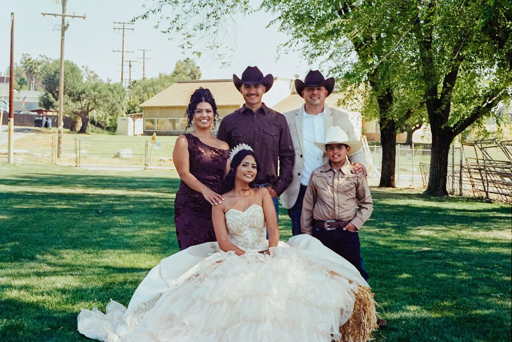 Group of five posing for a quinceañera photo outdoors, with the birthday girl in a white gown seated on a hay bale.