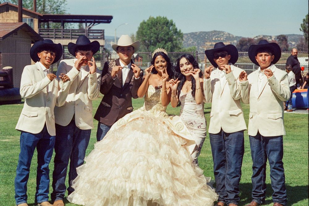 Young woman in elegant quinceañera ball gown poses with groomsmen in cowboy hats outdoors.