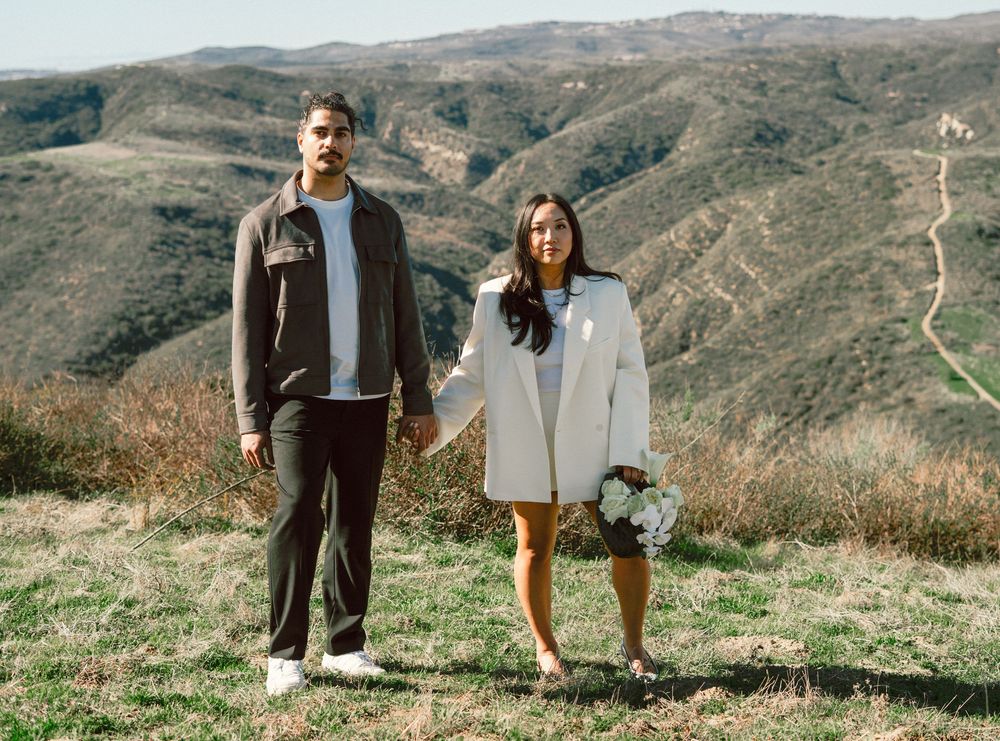 Engaged couple holding hands on a hilltop with scenic mountain landscape in background.