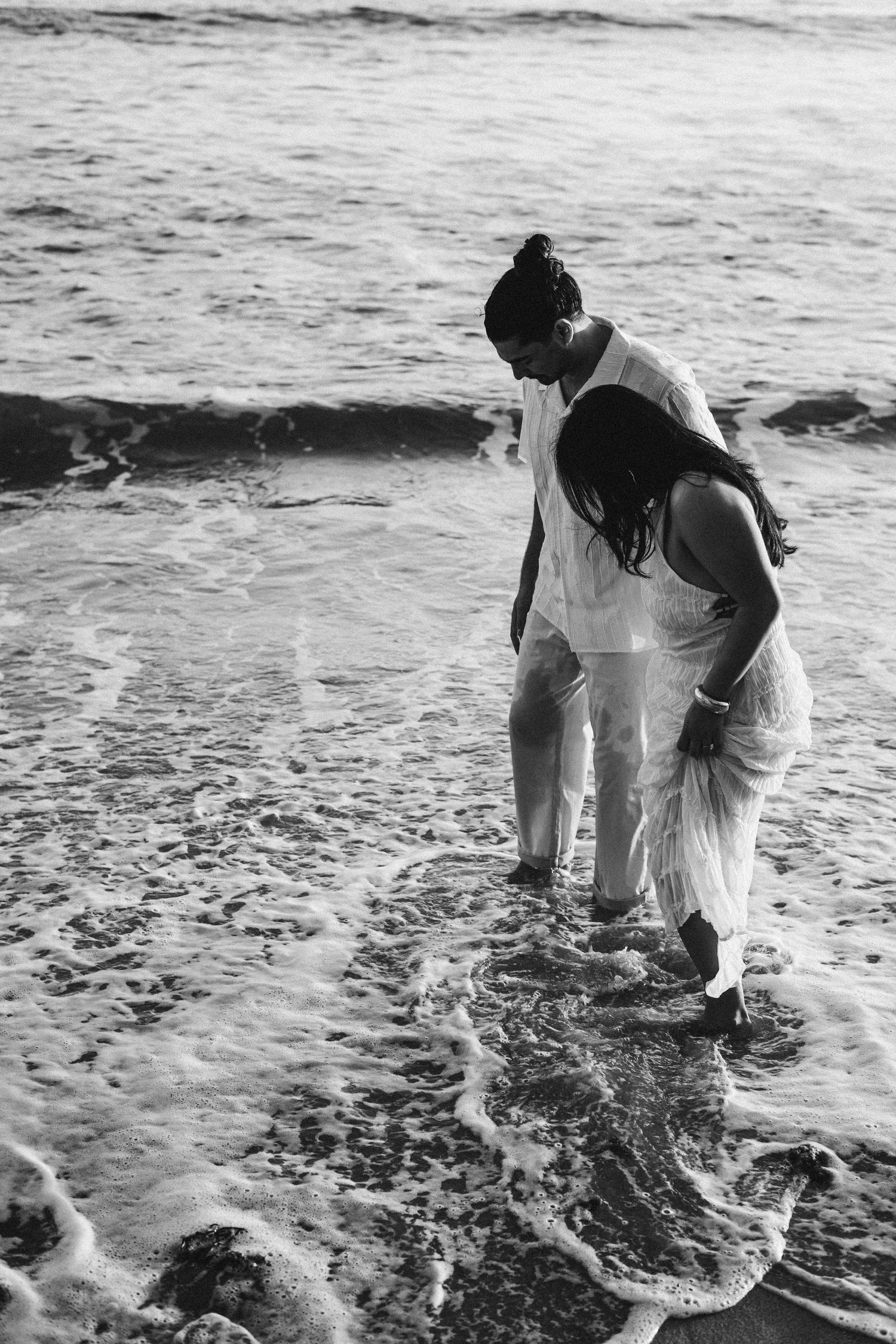 Black and white photo of couple wading together in ocean waves at the shoreline.
