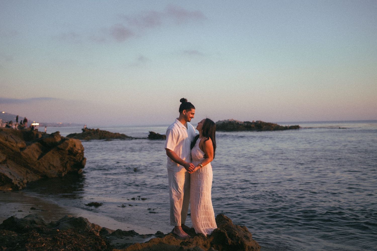 Couple standing face to face on coastal rocks during a romantic pastel sunset by the sea.