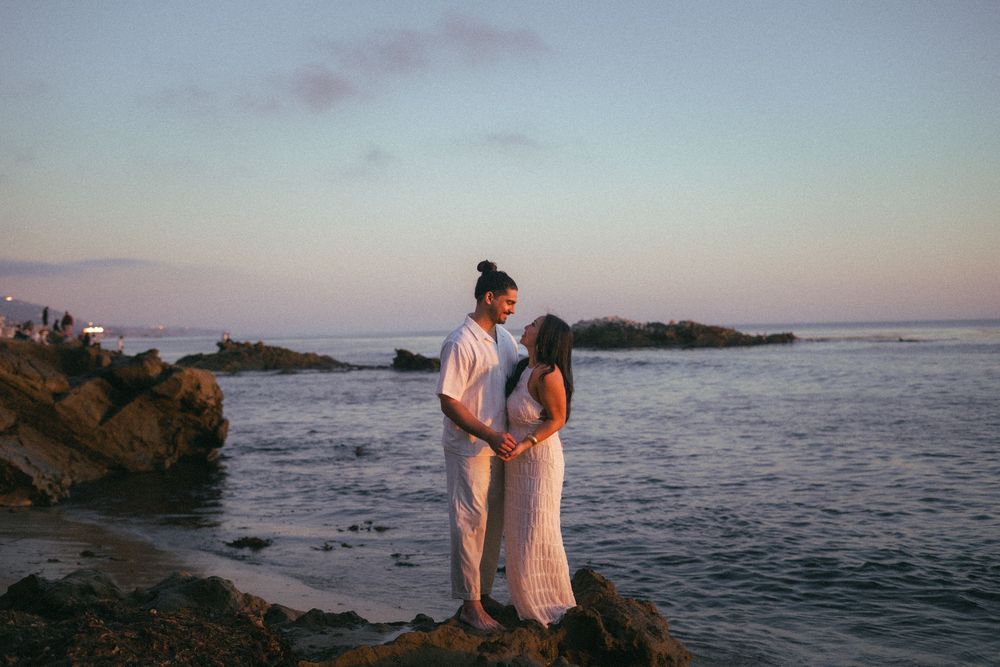Couple standing face to face on coastal rocks during a romantic pastel sunset by the sea.