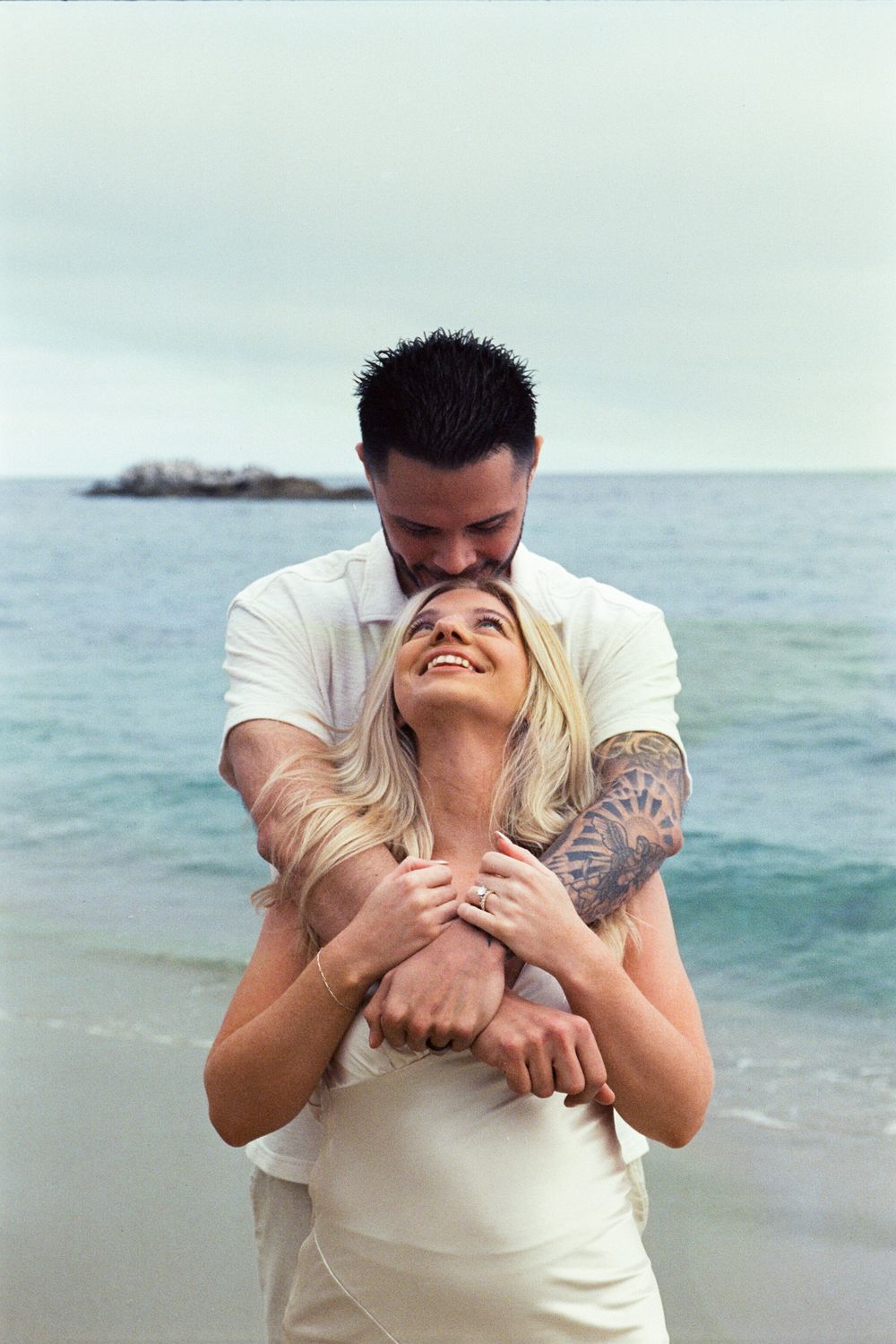 A couple embraces on a beach, the man kissing the woman's head as she smiles, both wearing white near the ocean.