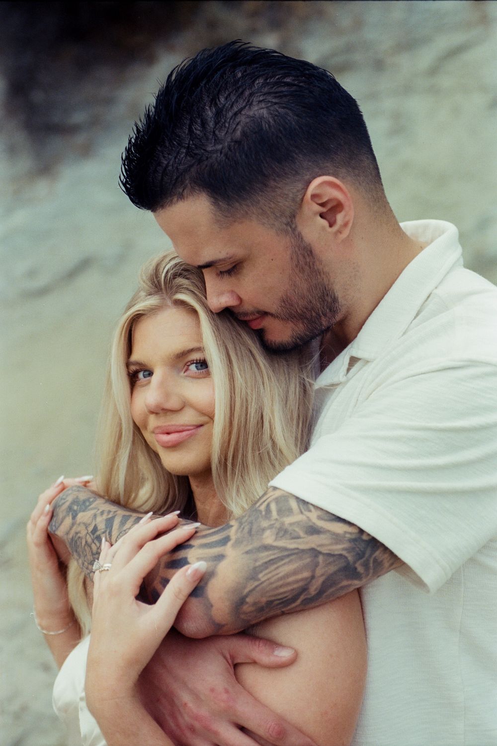 A tattooed man tenderly embraces a smiling blonde woman outdoors, both wearing white in a romantic couple portrait.