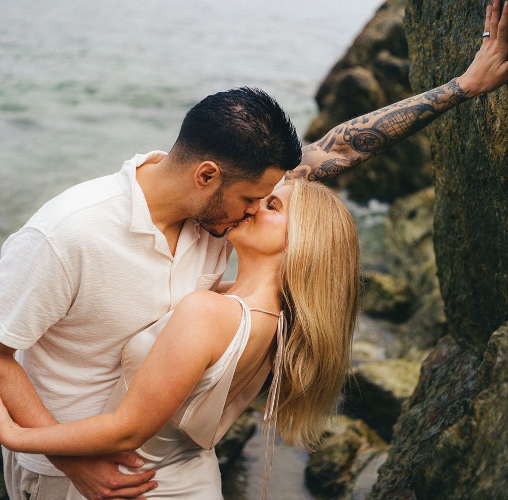 Couple sharing a romantic kiss by rocky shoreline, woman with blonde hair and tattoo, man in white shirt.