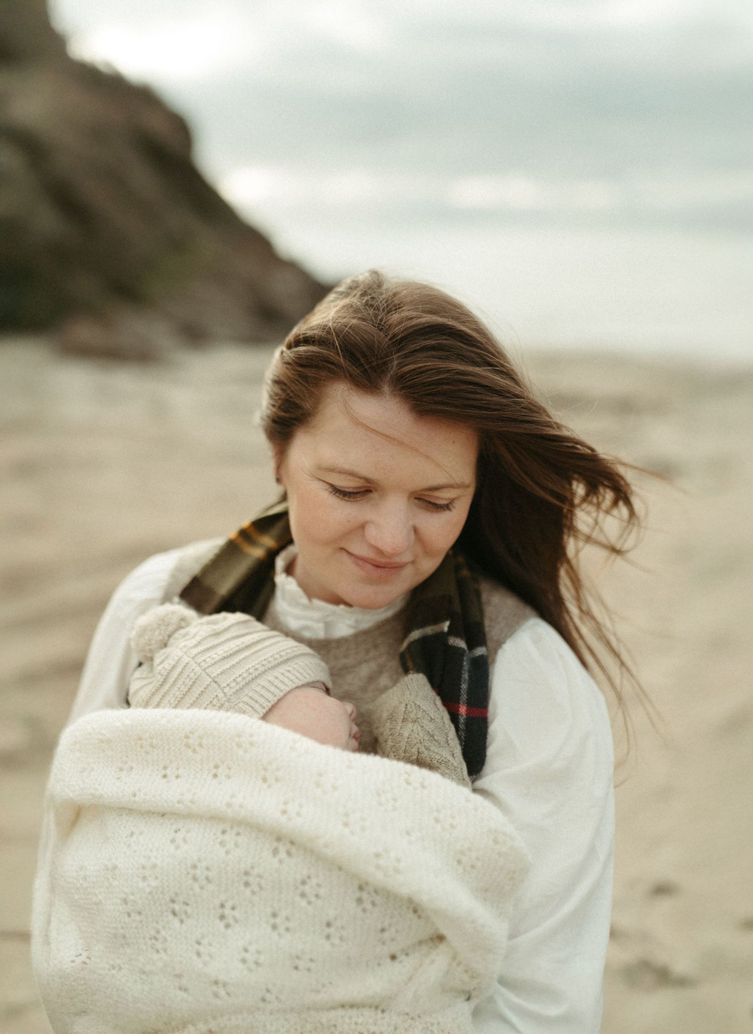 A person wrapped in a cozy cream knit blanket stands on a misty beach during an overcast day.