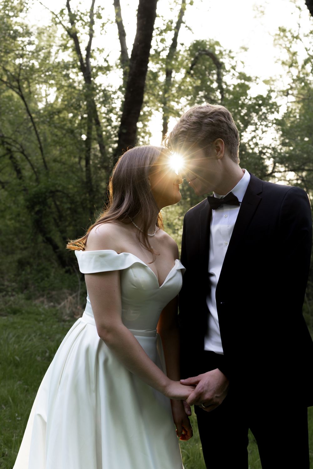 A couple shares an intimate moment in formal wedding attire against a backdrop of trees at sunset.