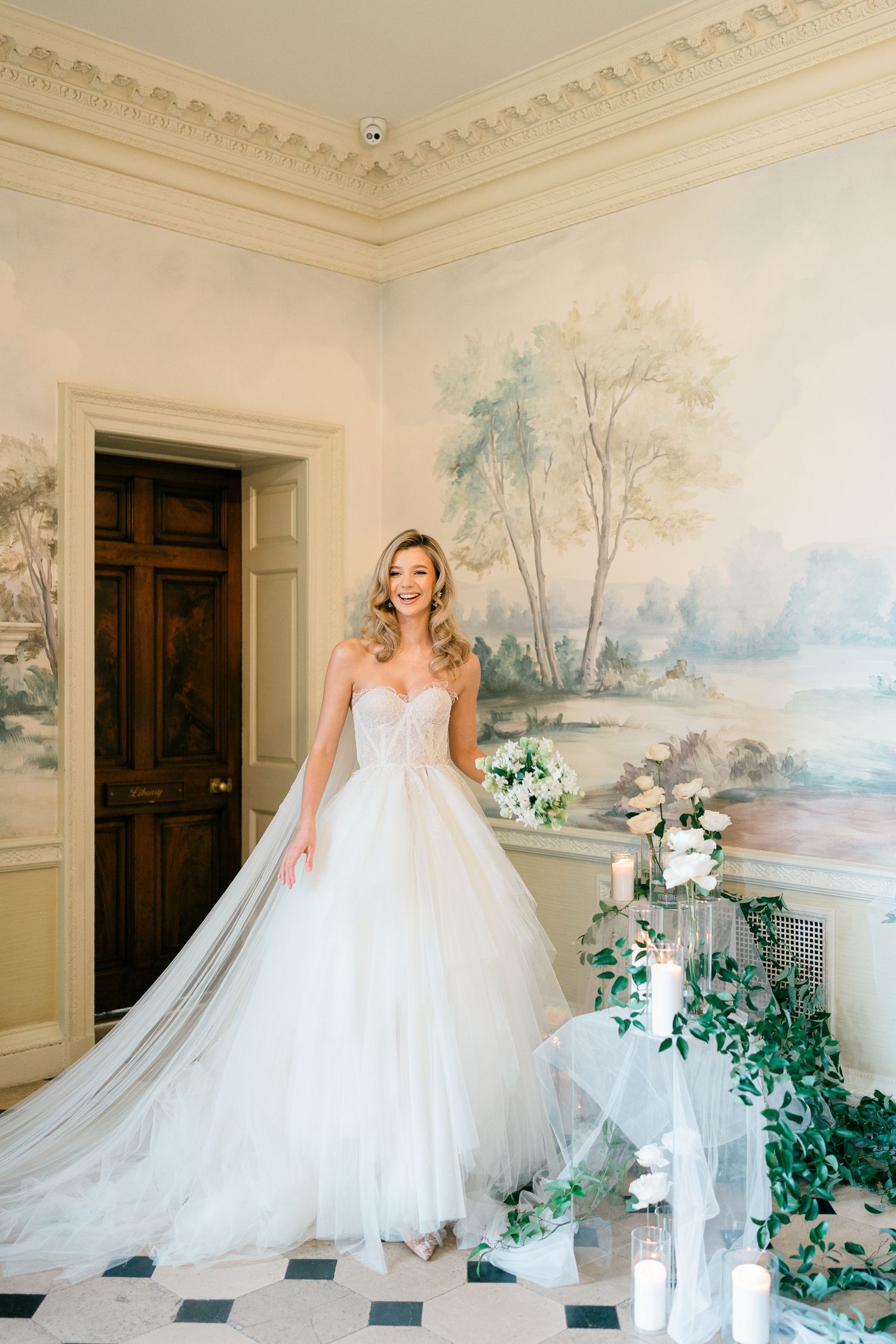 Elegant bride in flowing white ballgown poses in luxurious room with pastoral wall murals and ornate crown molding.