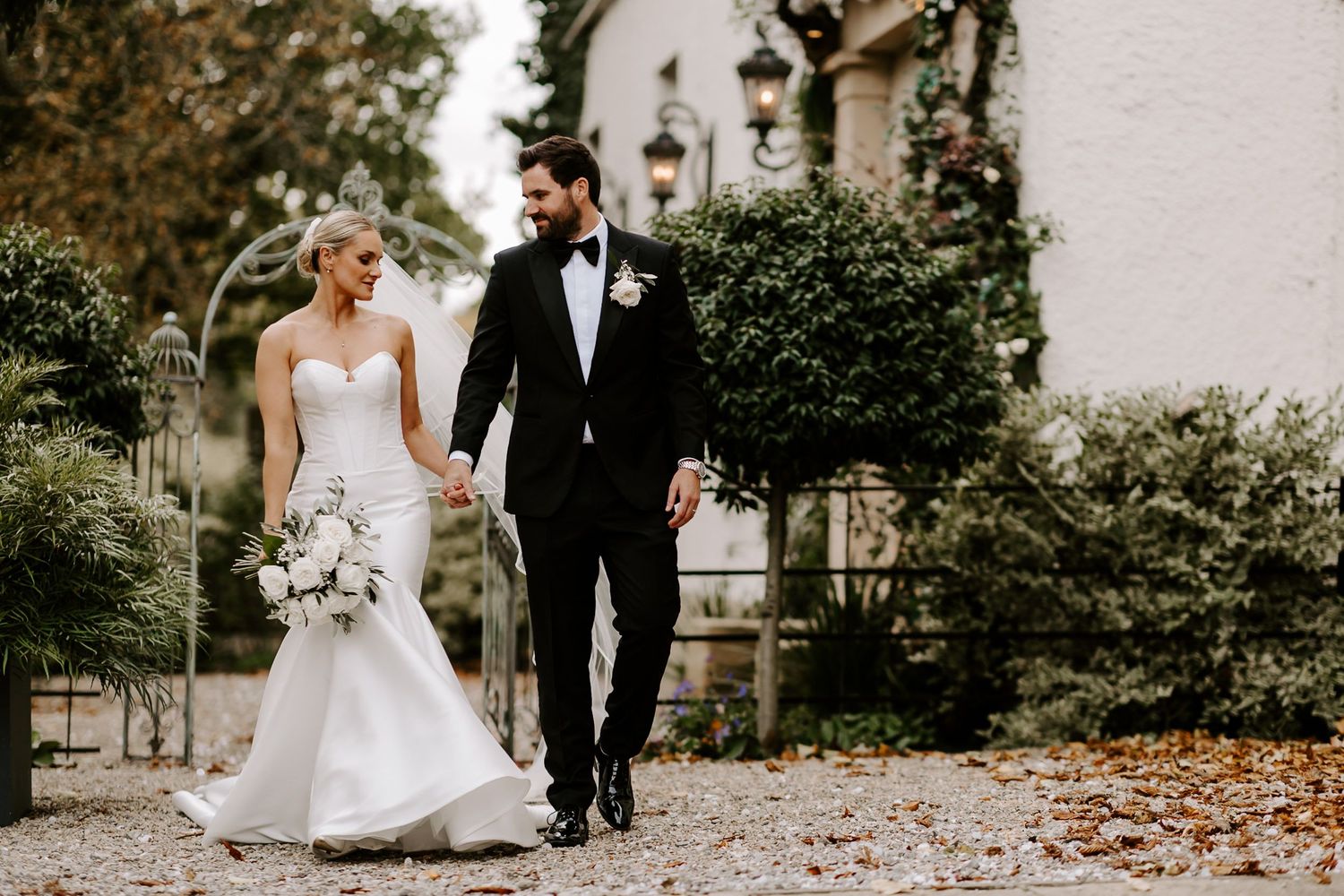 A couple in wedding attire walks together through a path covered in autumn leaves on their special day.