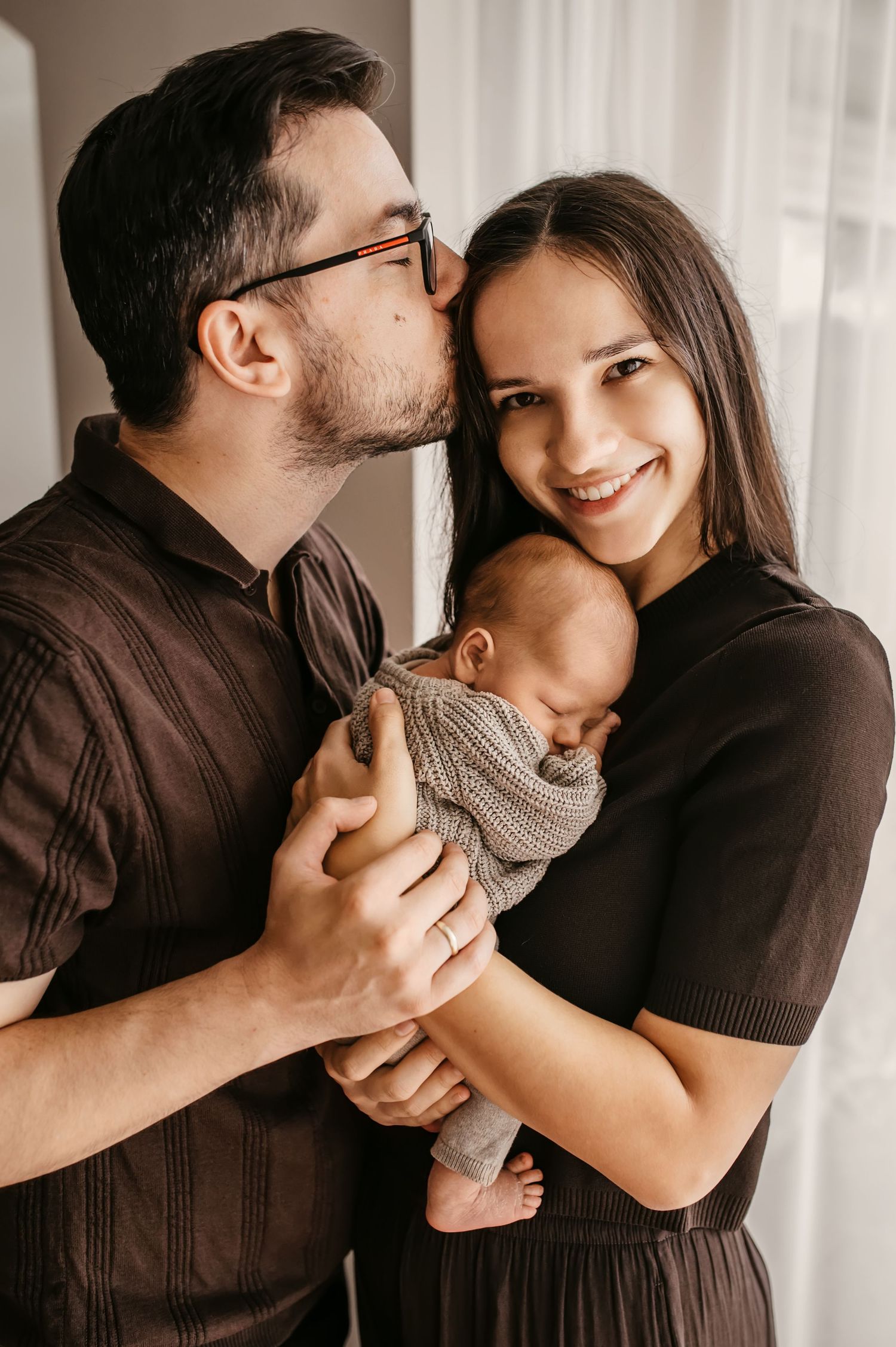 A couple shares a tender moment with their newborn baby wrapped in a gray knit blanket by a window.