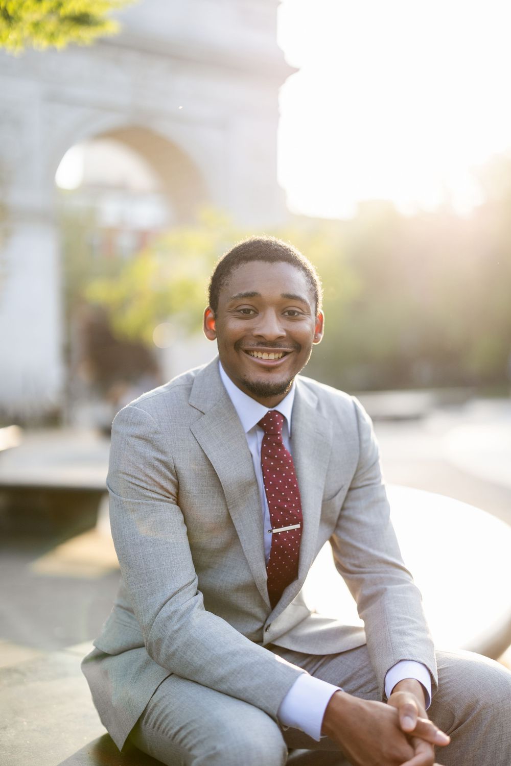 Professional in a light gray suit and red tie sits outdoors smiling during golden hour.