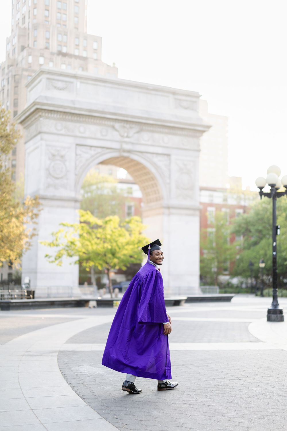 Graduate in purple cap and gown poses in front of Washington Square Arch on a sunny autumn day in New York City.