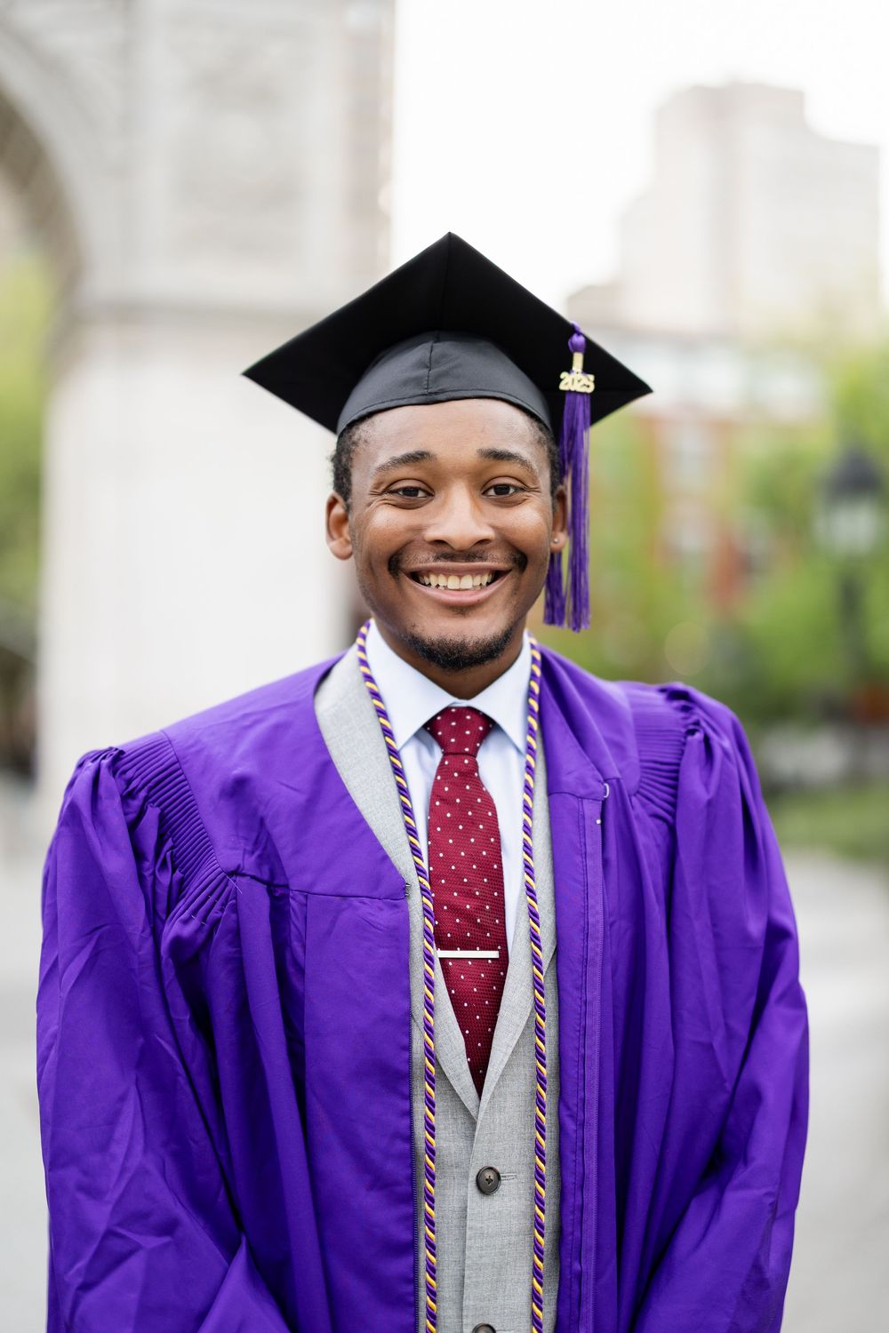 Graduate in purple cap and gown smiles brightly while wearing a grey suit and burgundy tie during commencement.