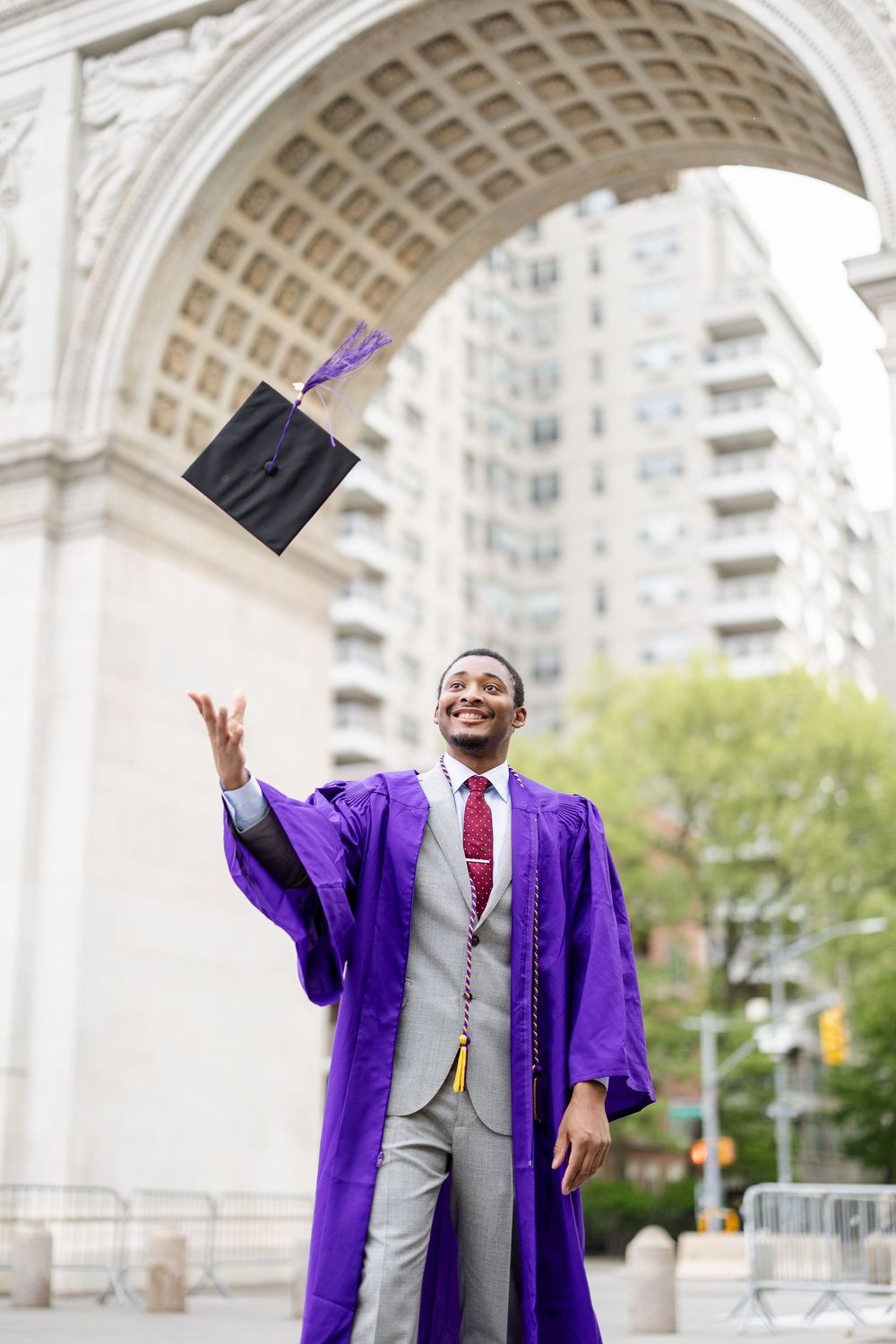 Graduate in purple regalia and grey suit celebrates with arms raised near stone archway in urban setting.