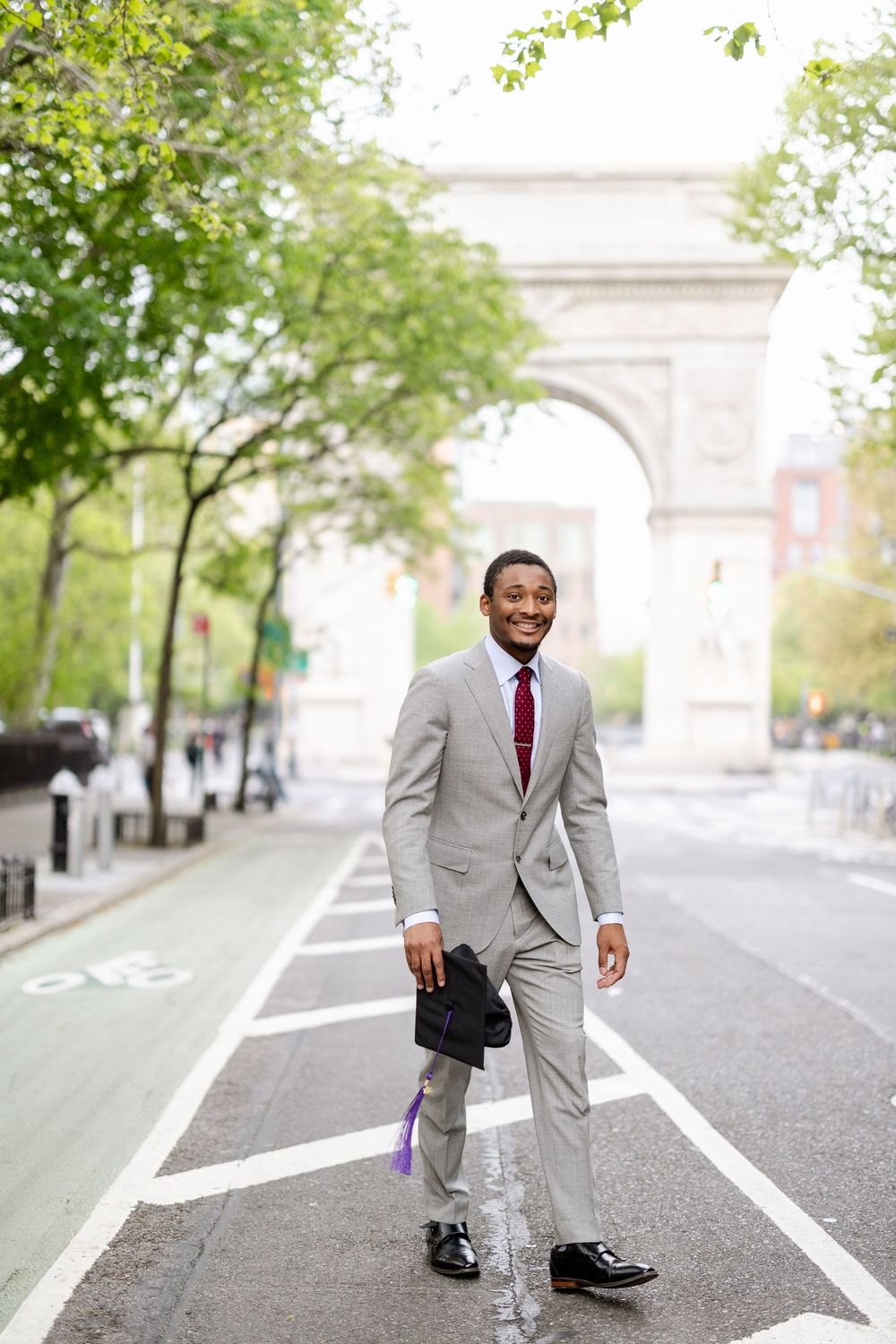 A person in a gray suit and red tie poses playfully near Washington Square Park arch in New York City.