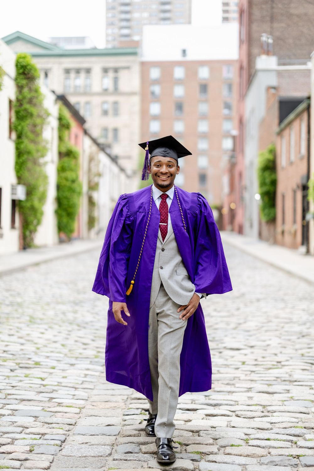 A graduate in purple regalia and gray suit poses on a cobblestone street for a series of celebratory photos.