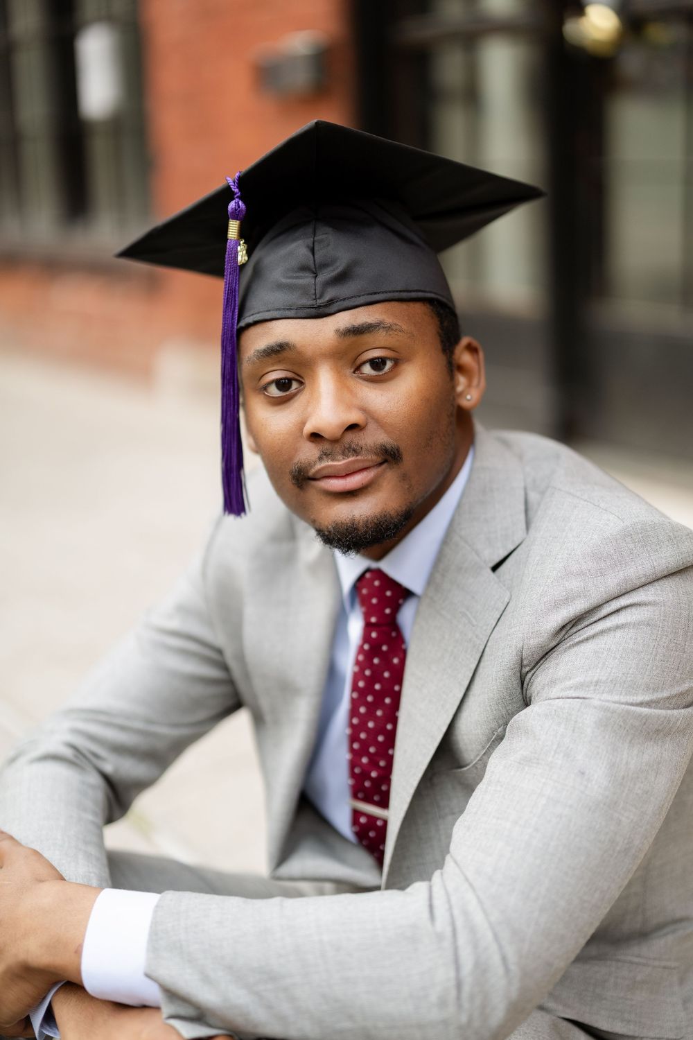 Graduate in light gray suit and burgundy polka dot tie poses with graduation cap in professional portrait series.