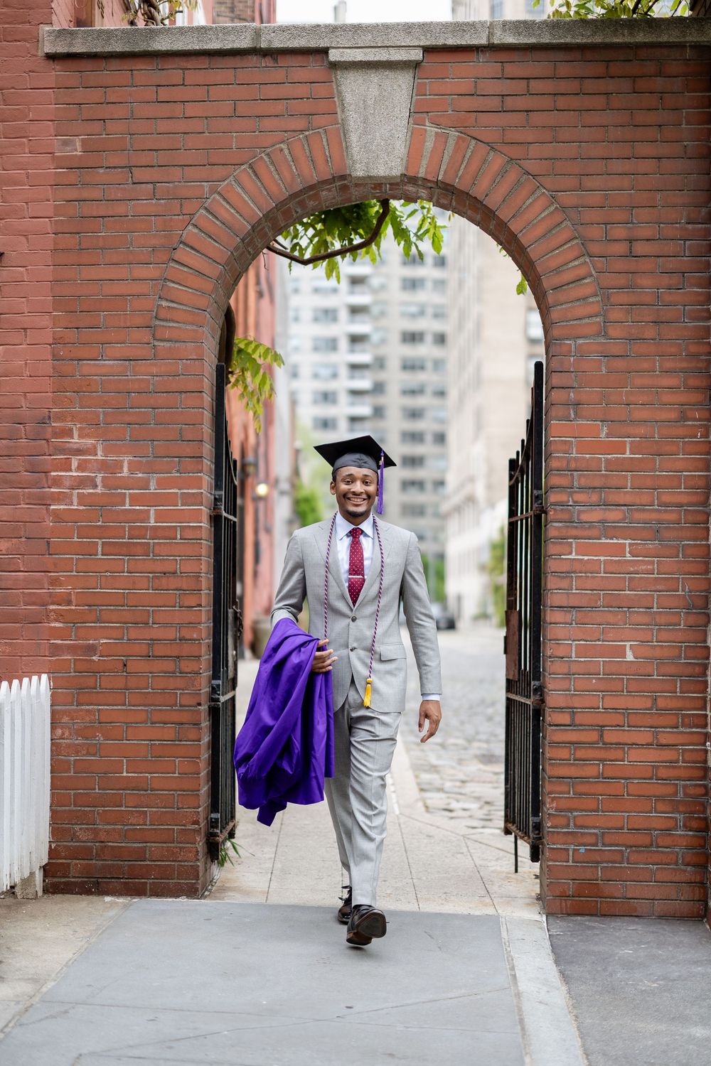 Graduate in grey suit walks through brick archways while carrying purple graduation gown.