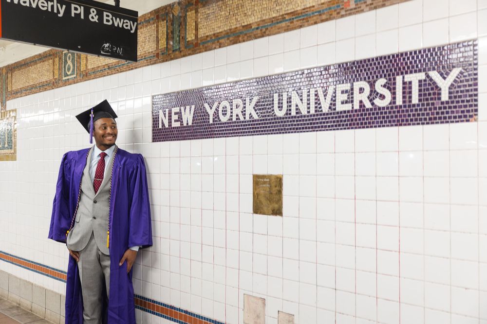 Graduate in purple academic regalia poses at New York University subway station platform with tiled wall.