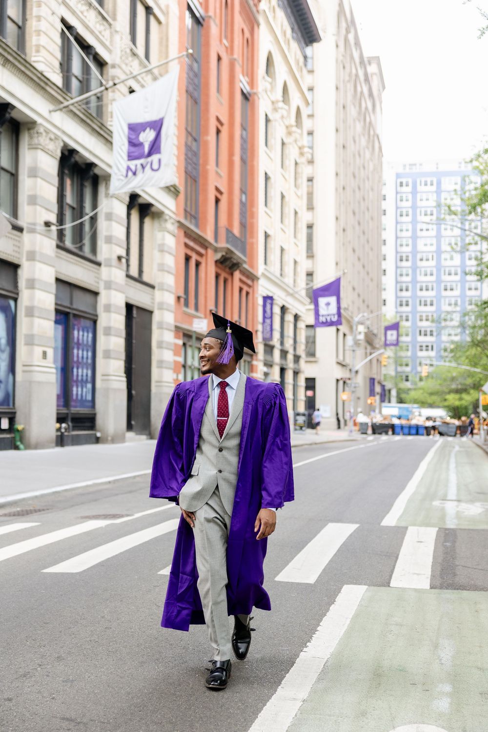 Graduate in purple commencement robe and gray suit walking confidently on a city street with NYU banners visible.