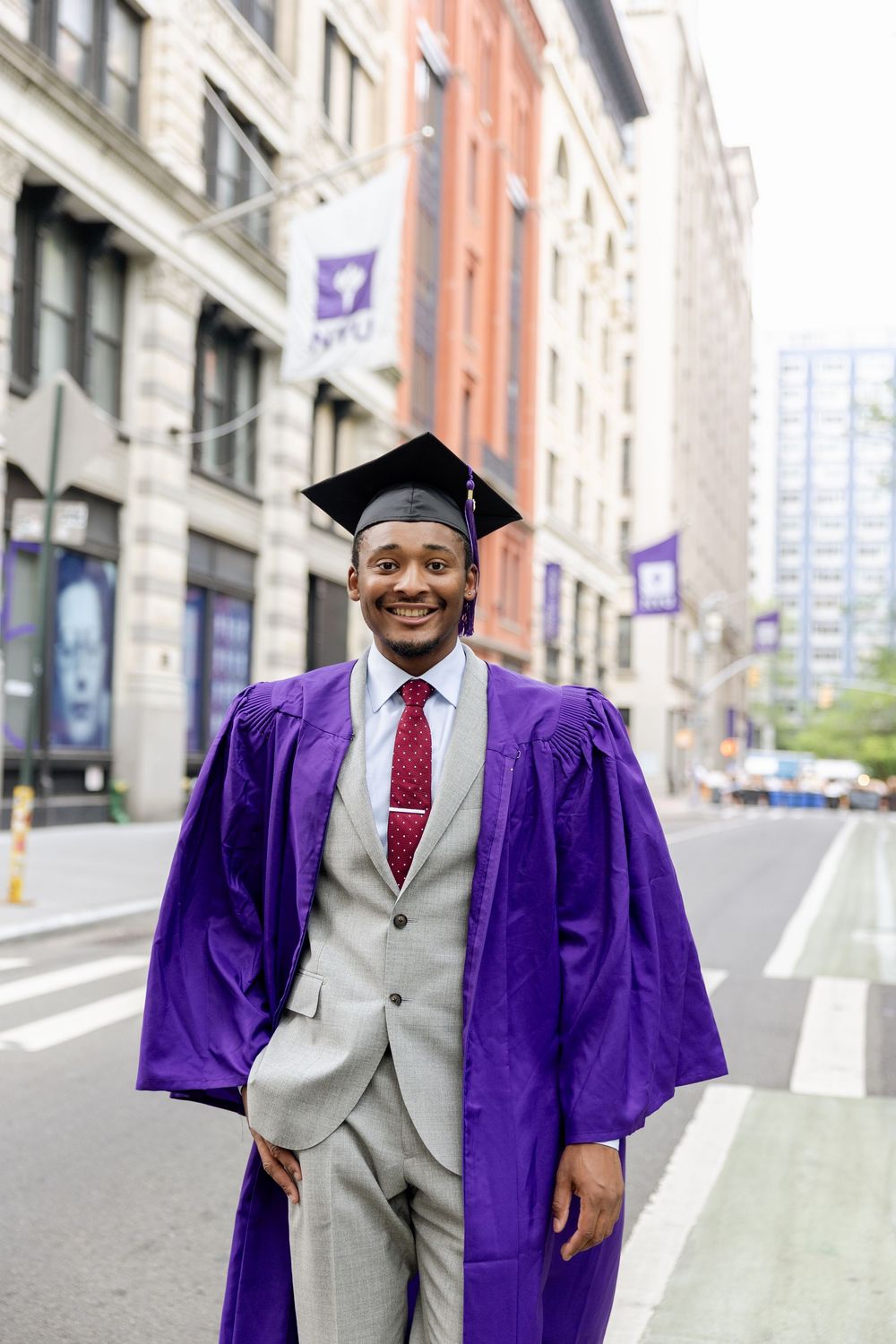 Graduate in purple NYU regalia and gray suit standing on urban street with university banners in background.