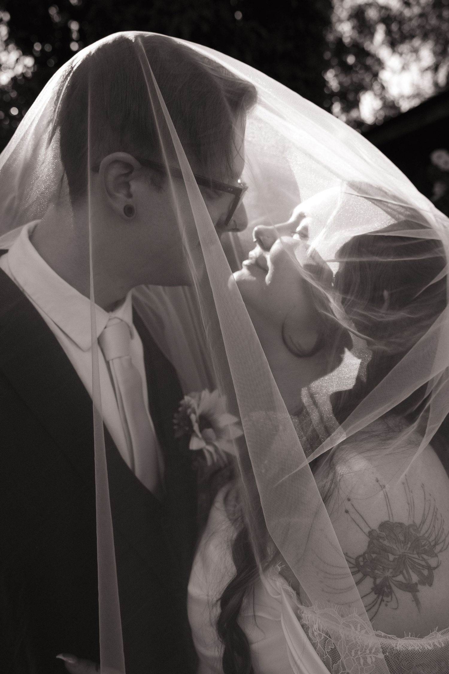 A romantic wedding kiss between a couple outdoors, with string lights and natural decor in the background.