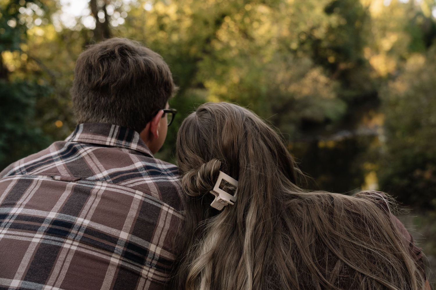 A couple sits together outdoors wearing casual clothing, viewed from behind against a sunlit forest backdrop.
