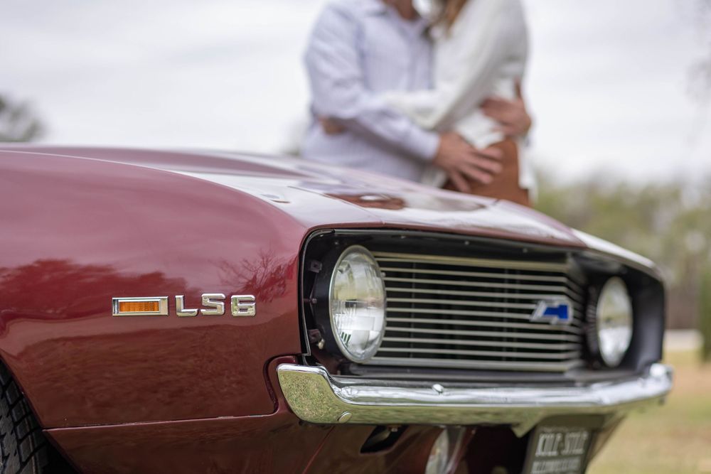 The front end of a classic car with a couple embracing in the background.