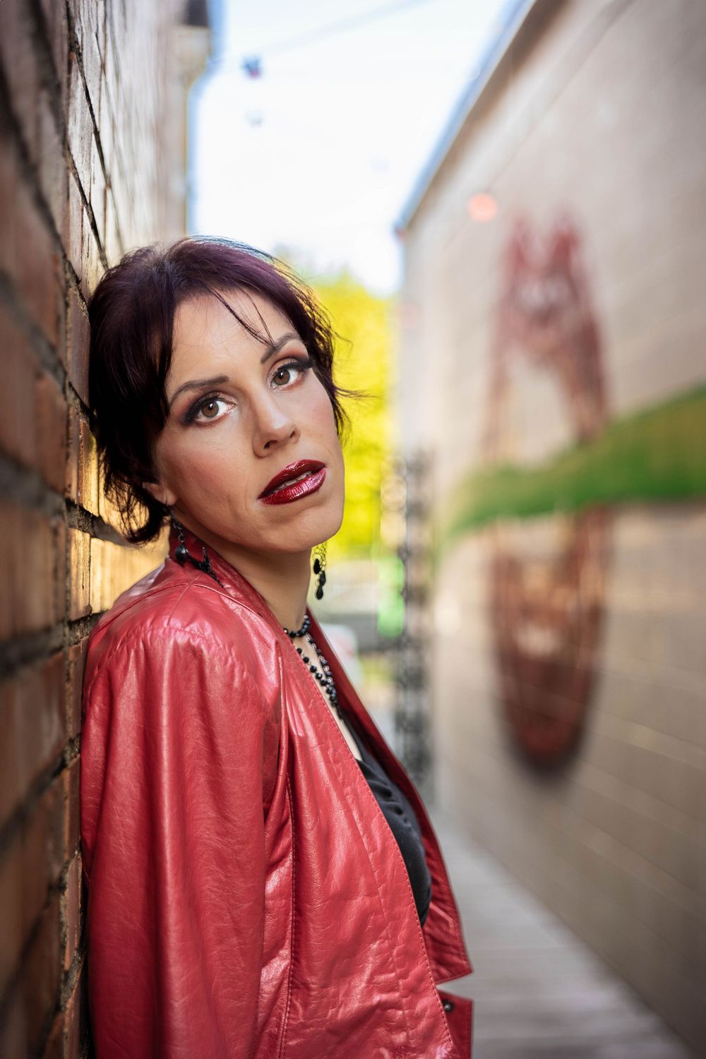 A woman in a red leather jacket poses in an alley with graffiti art.