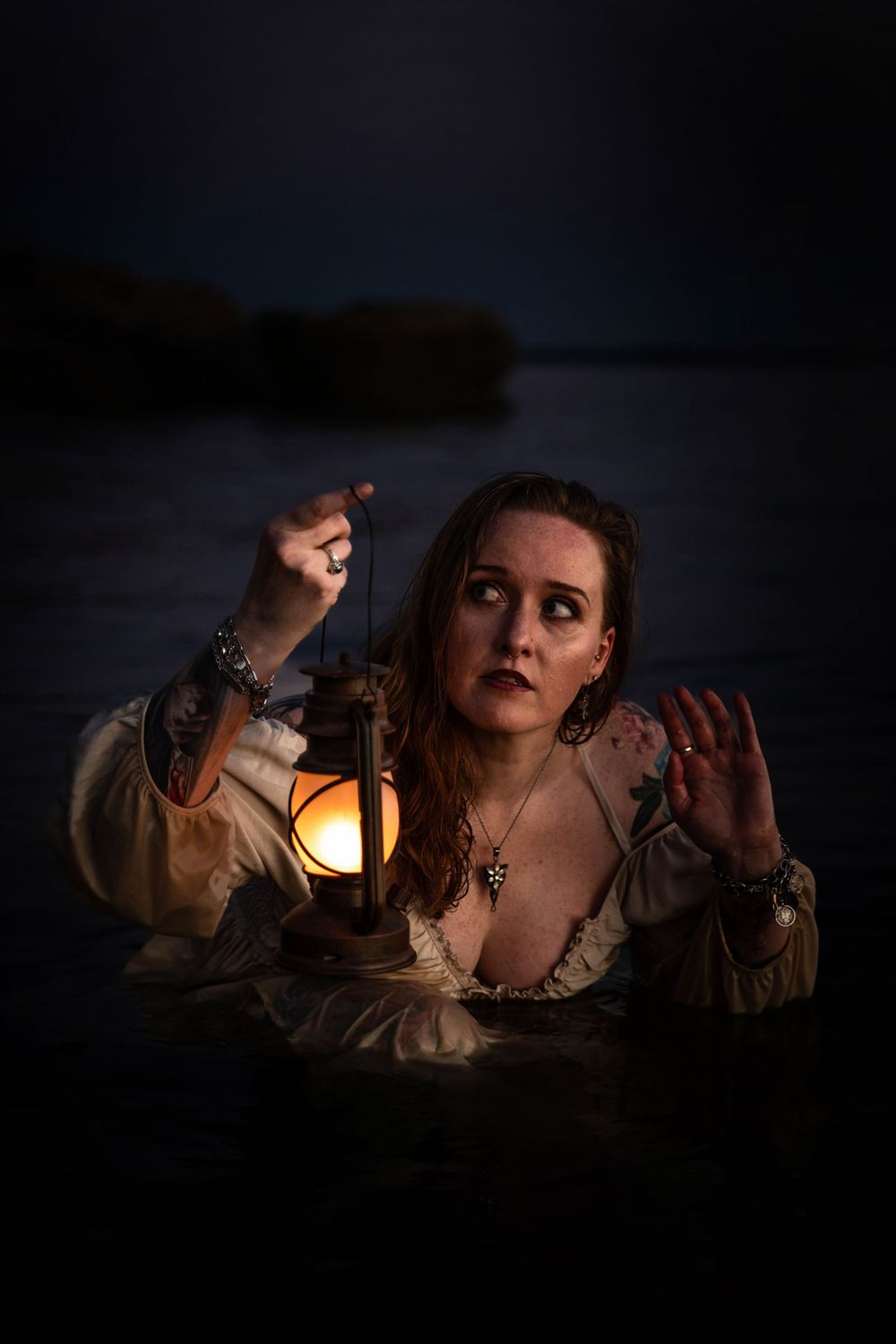 A woman wades through the water in a white dress at night time using lantern light.