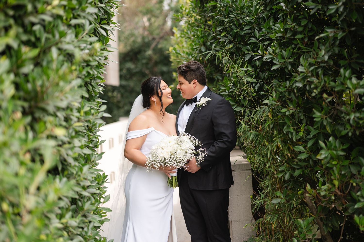 Bride and groom share romantic moments in a garden setting with lush greenery and white floral bouquet.