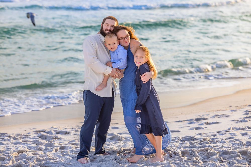 A family embraces while standing on a sandy beach with ocean waves and blue sky in the background.