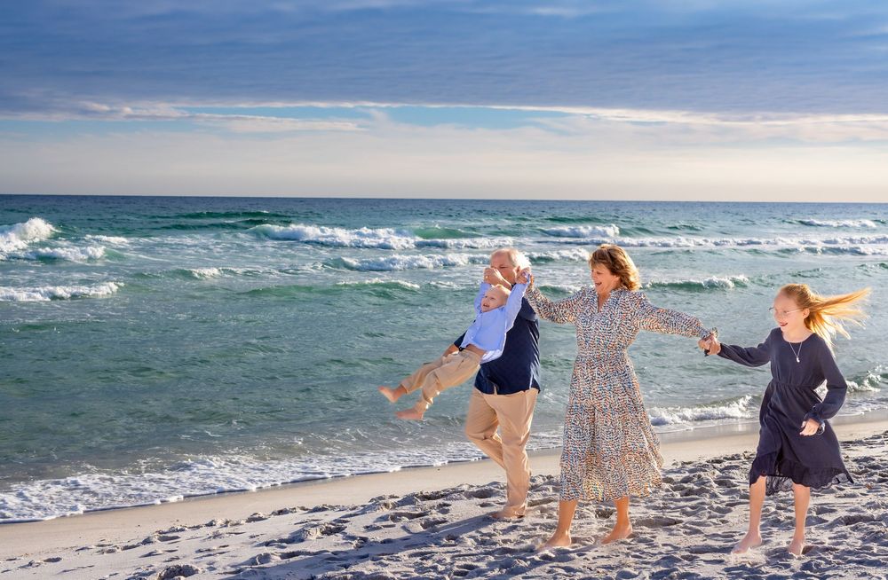 Three people hold hands and dance along the shoreline as waves crash on a beach during a cloudy day.
