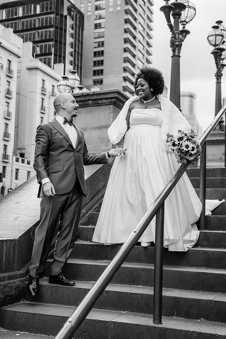 A black and white photo sequence showing a couple dancing playfully on urban stone steps near decorative lampposts.
