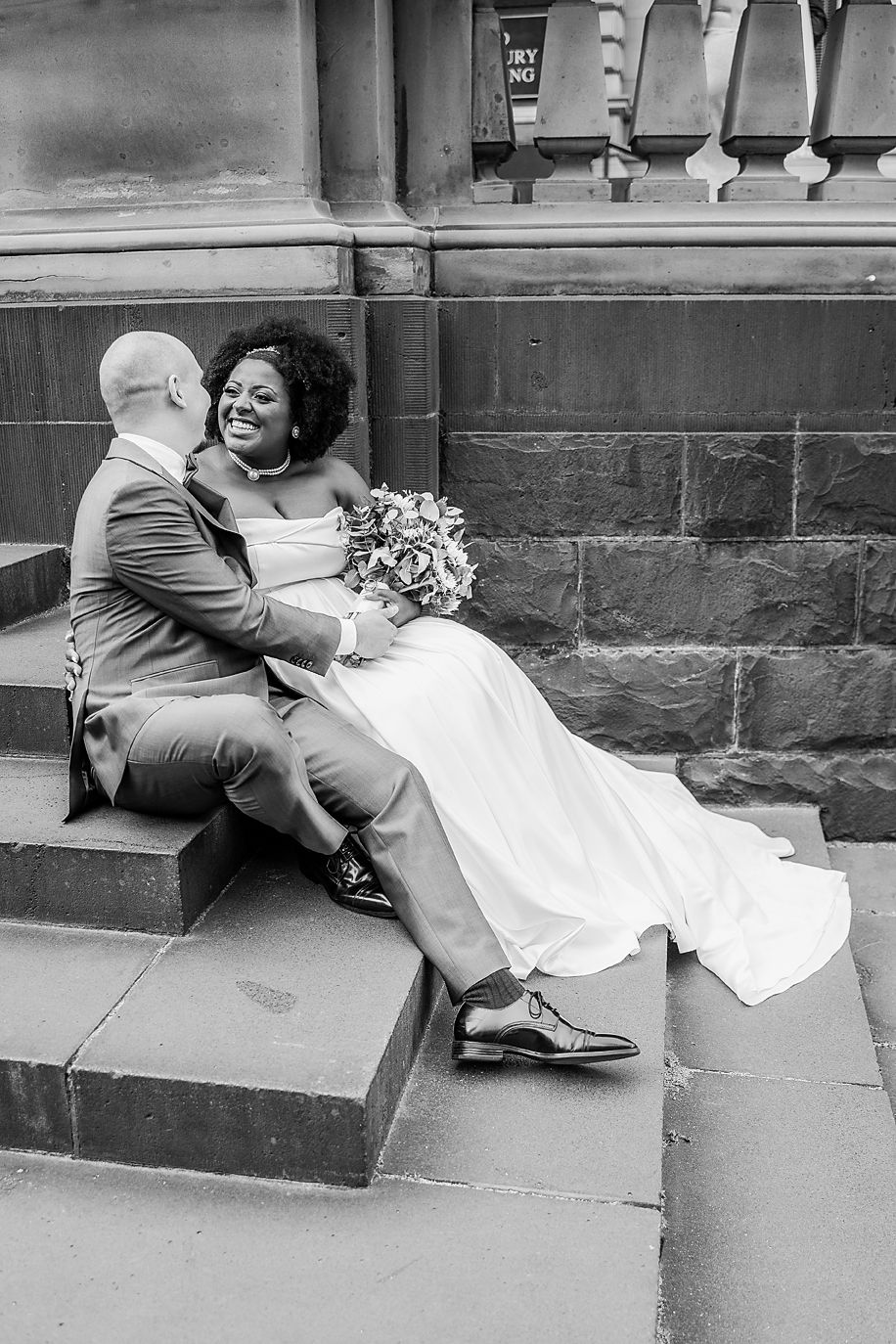 A couple in wedding attire sits playfully on stone steps in a series of black and white candid photos.