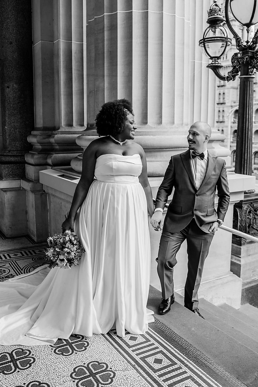 Black and white wedding photo showing a couple posing between elegant columns with decorative floor patterns.