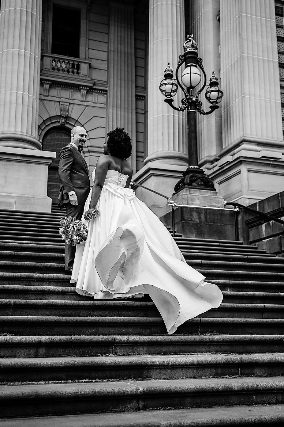 A sequence of black and white photos showing a bride in a flowing dress jumping down stone steps in front of a classical building.