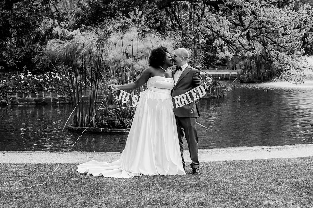 A couple in wedding attire poses near a scenic lake surrounded by lush greenery and trees.