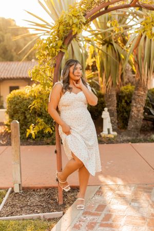 A person in a white sundress poses under a floral arch with palm trees and golden sunlight in the background.
