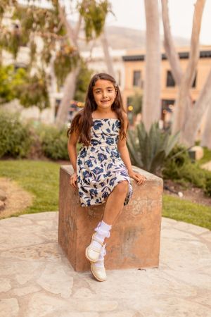 A young person in a floral dress sits on a stone block in a garden setting.
