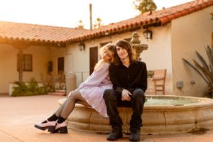 A couple sits intimately together on a stone fountain ledge at sunset near a Spanish colonial style building.