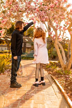 Two figures admire blooming cherry blossom trees along a brick pathway at golden hour.