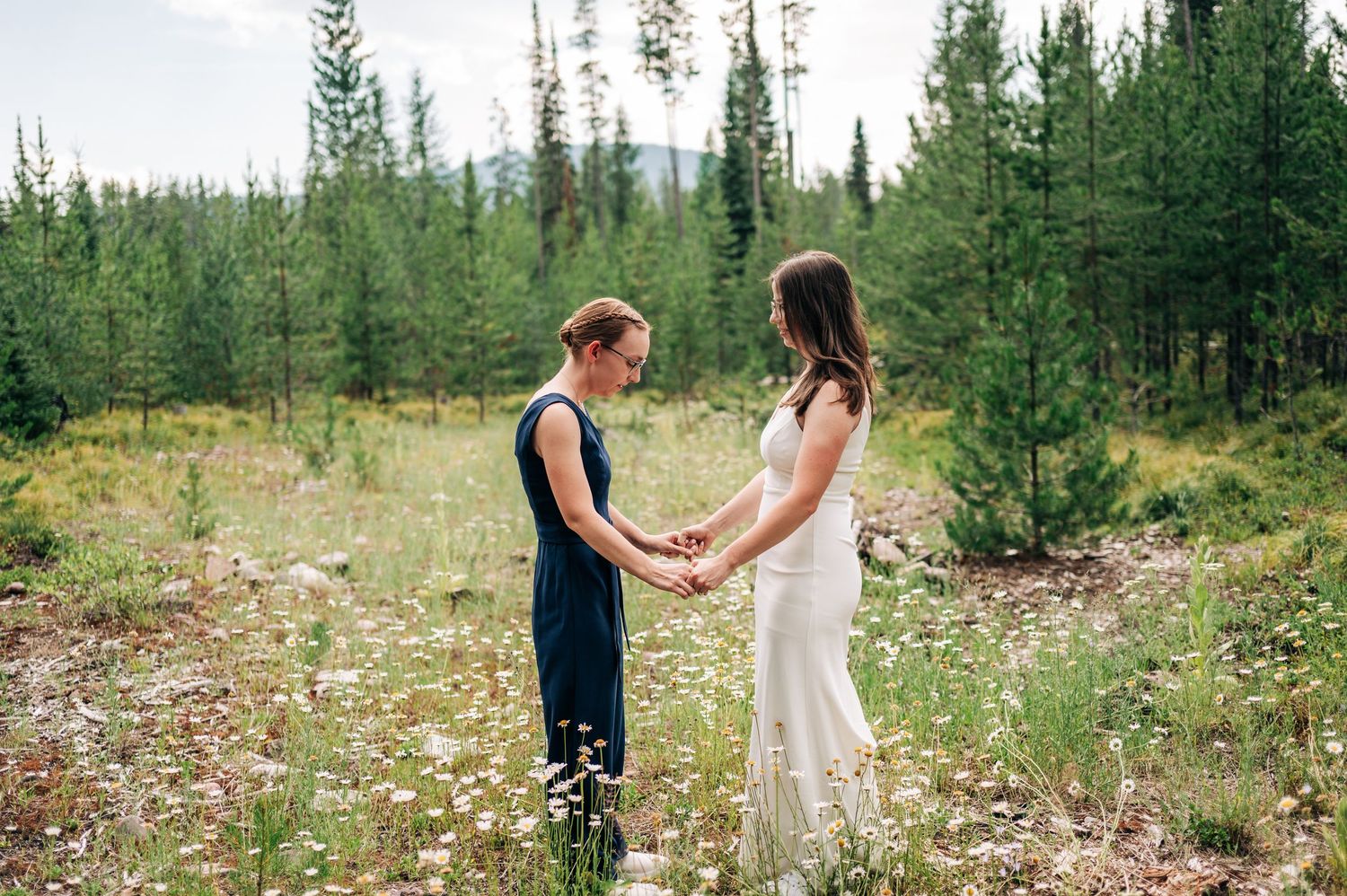 A couple in wedding attire walks through a serene forest setting surrounded by evergreen trees and wildflowers.