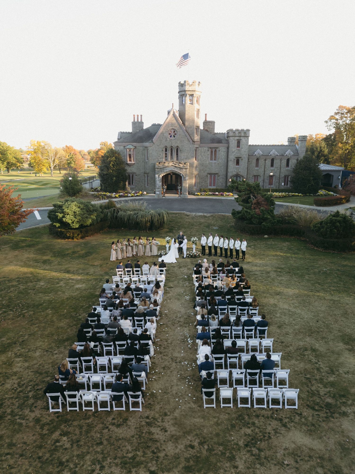 Aerial view of a castle-like venue with white chairs arranged in rows on the lawn for an outdoor wedding ceremony.